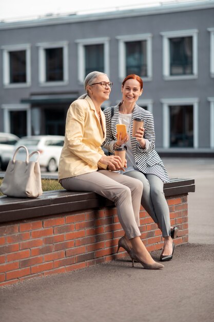 Aproveitando o tempo livre. Duas mulheres modernas sentadas no banco ao ar livre com copos de papel com café e um smartphone enquanto sorriem
