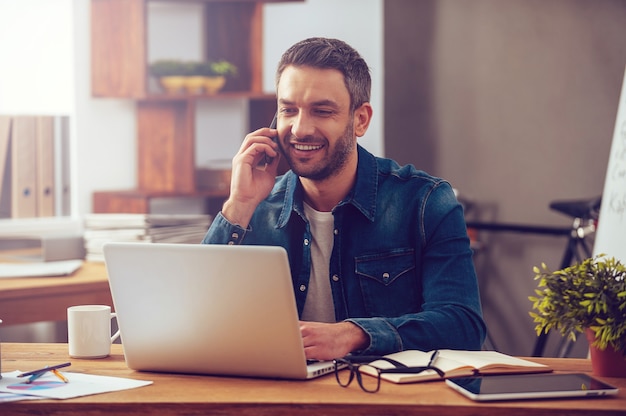 Aproveitando um bom dia de trabalho. Jovem confiante trabalhando em um laptop e falando ao telefone celular enquanto está sentado em seu local de trabalho no escritório