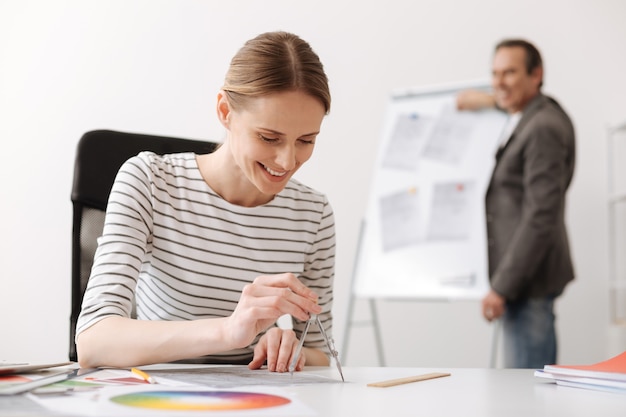 Aproveite seu trabalho. engenheira profissional alegre sentada à mesa e usando bússolas enquanto seu colega fica de pé ao fundo