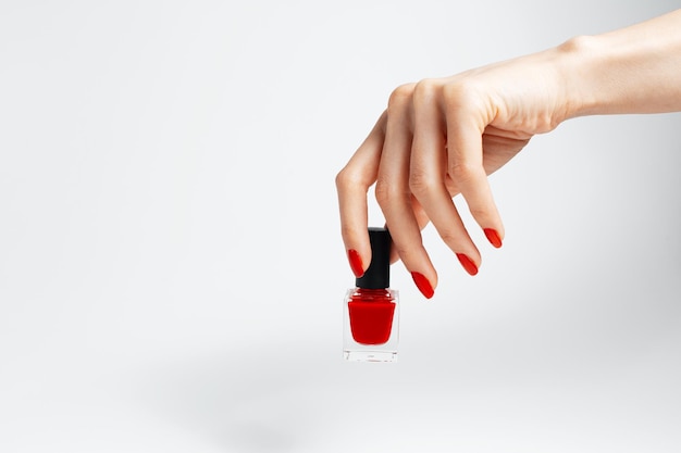 Aproximação da mão feminina com manicure vermelha segurando um esmalte brilhante de cor vermelha sobre fundo branco