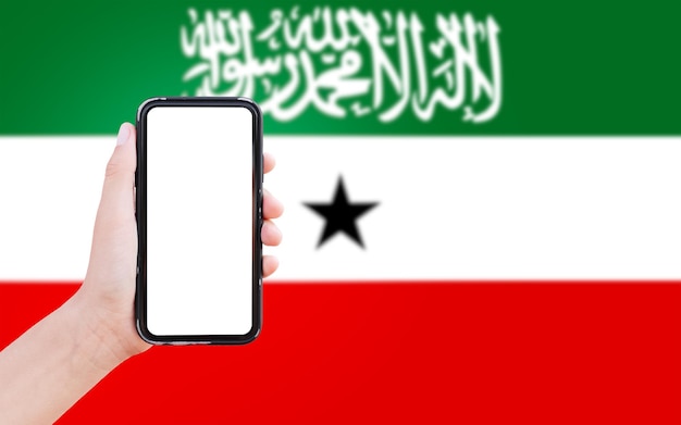 Aproximação da mão masculina segurando smartphone com espaço em branco na tela no fundo da bandeira desfocada da somalilândia