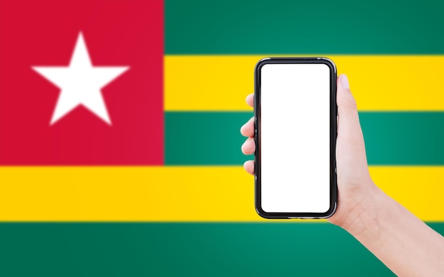 Aproximação da mão masculina segurando smartphone com espaço em branco na tela no fundo da bandeira desfocada do Togo