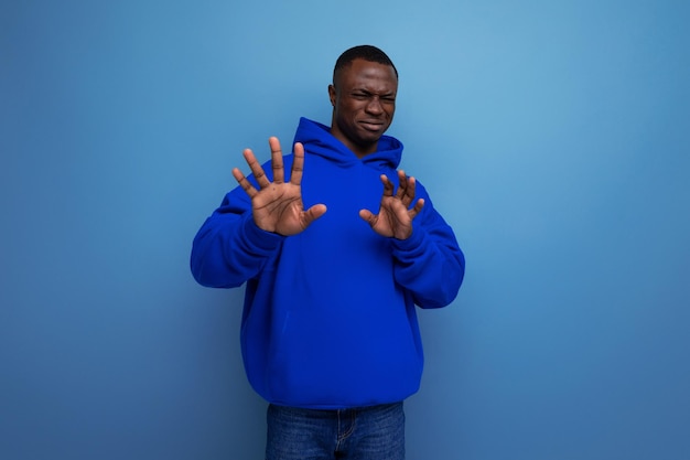 Aproximação de um jovem africano emocional em um moletom azul com capuz em um fundo de estúdio