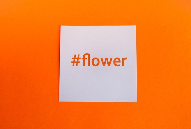 Aproximação de uma flor da palavra flor com uma hashtag em um pedaço de papel quadrado branco em um plano de fundo laranja