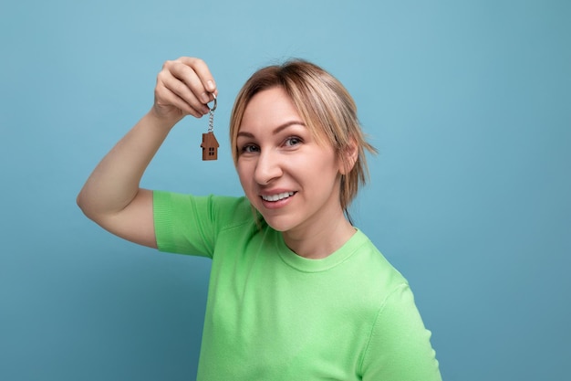 Aproximação de uma loira bonita e sortuda com aparência casual segurando as chaves do apartamento em fundo azul