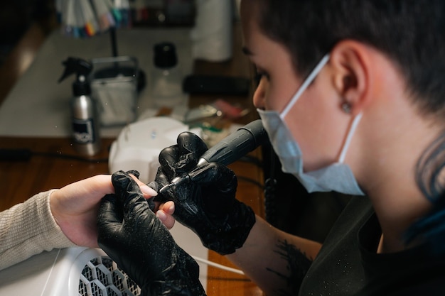 Aproximação do mestre feminino irreconhecível em máscara protetora polindo o ponto de corte da cutícula nas unhas com máquina de manicure elétrica