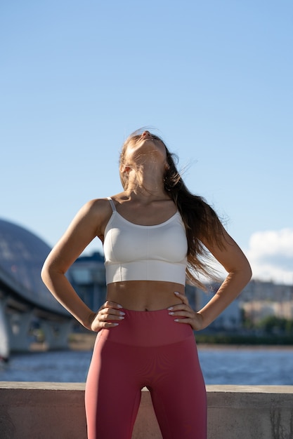 Apta a jovem mulher atlética, alongando o corpo no aterro. fitness feminino em legging rosa, fazendo exercícios de aquecimento para os músculos do pescoço ao ar livre.