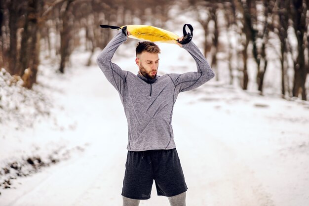 Apta o esportista fazendo exercícios com pesos em pé no caminho de neve na natureza no inverno. Fitness de inverno, musculação, hábitos saudáveis