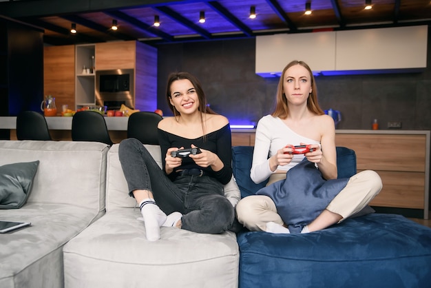 Apuestas alegres jóvenes amigas sentadas en el sofá en el apartamento contemporáneo y disfrutando de videojuegos