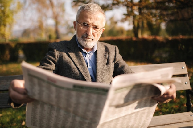 El apuesto abuelo con una hermosa barba en una chaqueta gris se sienta en un banco en el parque y lee un periódico.