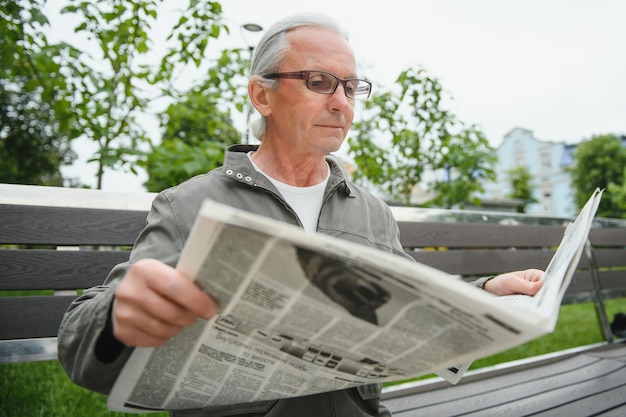 Apuesto abuelo se sienta en un banco en el parque y lee un periódico