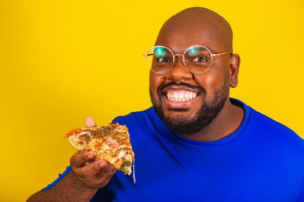 Un apuesto afrobrasileño con anteojos, camisa azul sobre fondo amarillo, sosteniendo una porción de pizza y sonriendo