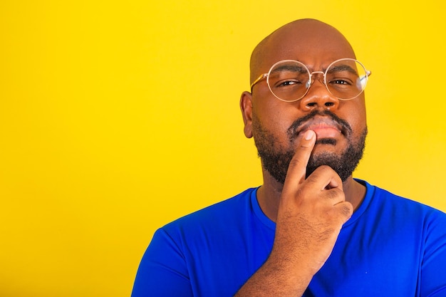 Un apuesto afrobrasileño con gafas de camisa azul sobre fondo amarillo con las manos en la barbilla cuestionando la reflexión pensando pensando eligiendo razonamiento