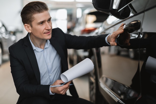 Apuesto agente de seguros de automóviles sentado cerca de un auto nuevo haciendo un chequeo tomando notas en el automóvil