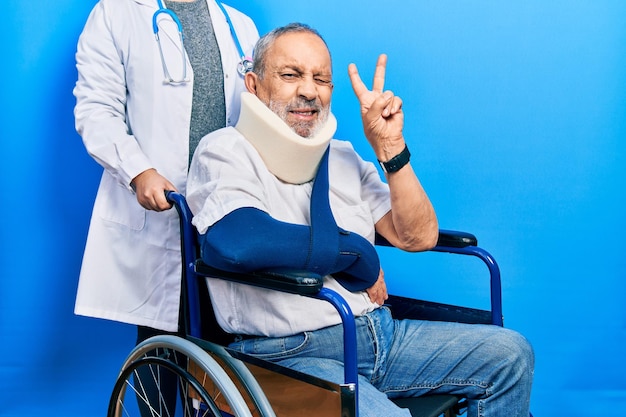 Un apuesto anciano con barba sentado en silla de ruedas con cuello sonriendo con cara feliz guiñando un ojo a la cámara haciendo el signo de la victoria con los dedos. número dos.