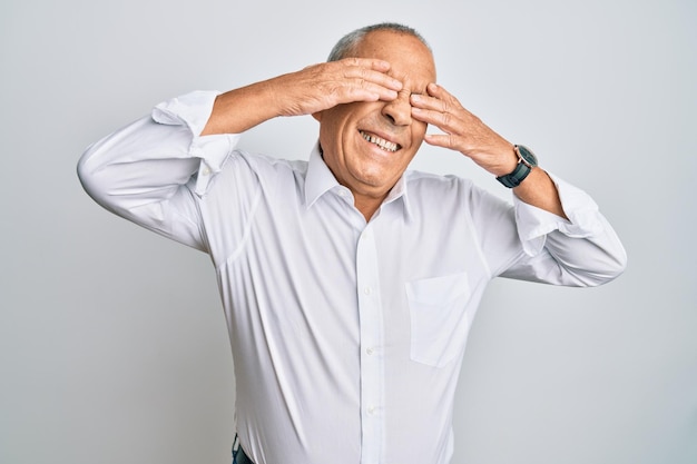 Apuesto anciano con camisa blanca casual que cubre los ojos con las manos sonriendo alegre y divertido concepto ciego