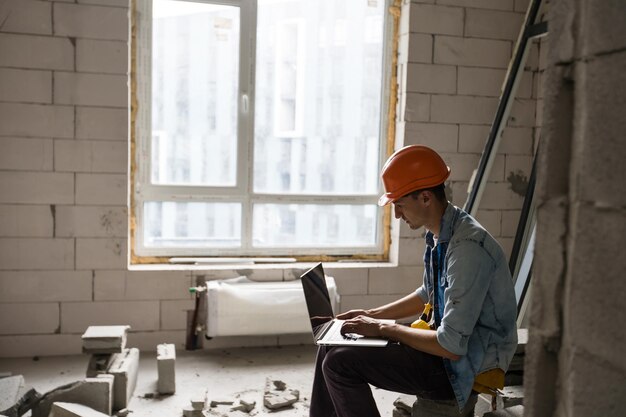 Apuesto arquitecto o capataz en casco trabajando con computadora portátil en los dibujos del apartamento en el interior de la estructura blanca