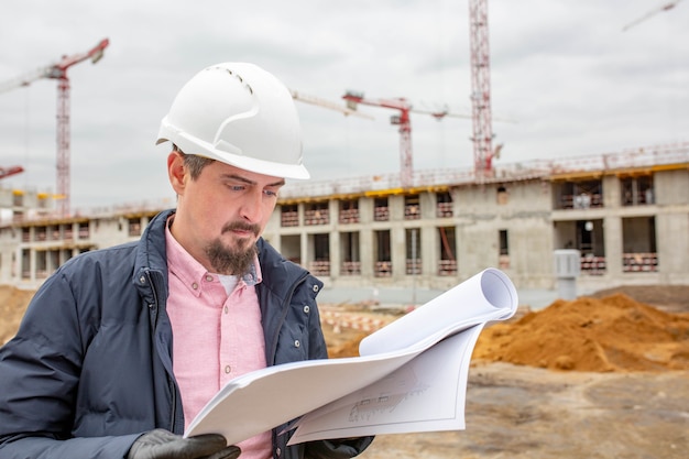 Apuesto arquitecto o supervisor de pie al aire libre en un sitio de construcción sosteniendo un plano en sus manos