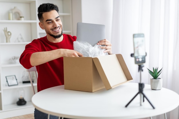 Apuesto bloguero árabe unboxing paquete con laptop en cámara