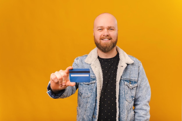 Un apuesto calvo barbudo que sostiene una tarjeta de crédito está sonriendo a la cámara