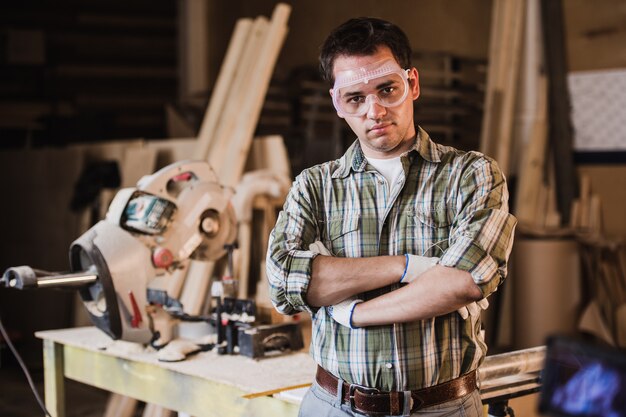 Apuesto carpintero con gafas protectoras está mirando a la cámara y sonriendo mientras está de pie cerca de su objeto de madera en el taller