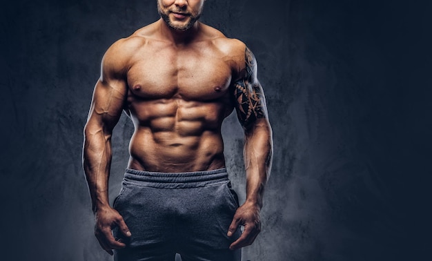 Un apuesto culturista sin camisa con un elegante corte de pelo y barba, con un tatuaje en el brazo, posando en un estudio. Aislado en un fondo oscuro.