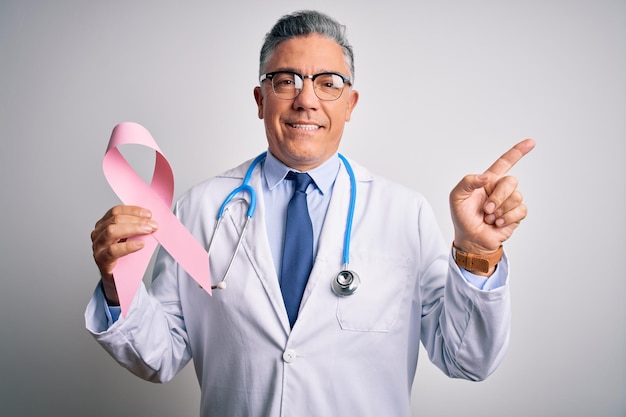 Un apuesto doctor canoso de mediana edad que sostiene una cinta rosa de cáncer muy feliz señalando con la mano y el dedo hacia un lado