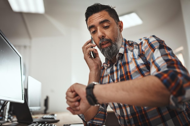 Un apuesto empresario exitoso está hablando por un teléfono celular mientras trabaja en una oficina. Él está mirando su reloj para no llegar tarde.