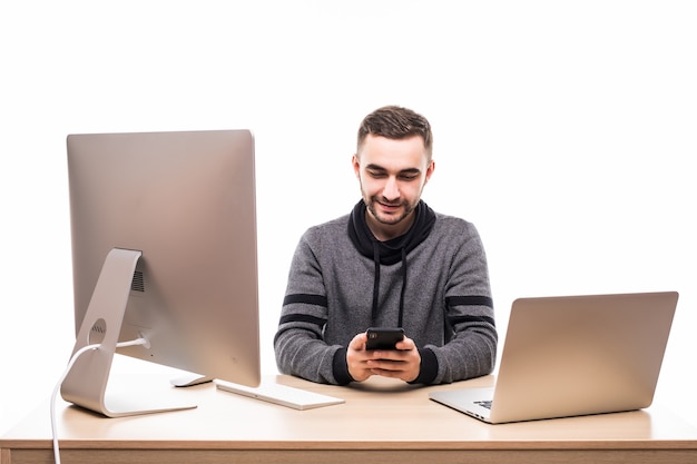 Apuesto empresario sentado detrás de su escritorio con computadora portátil y pc y escribiendo en el teléfono móvil aislado en blanco