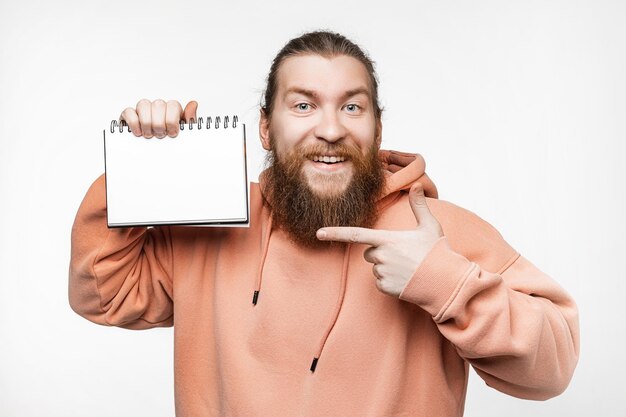 Un apuesto escandinavo feliz sosteniendo un bloc de notas y apuntando a una maqueta de papel aislada en un fondo gris Chico con peinado de jengibre y barba en una sudadera Chico sosteniendo un cuaderno para publicidad