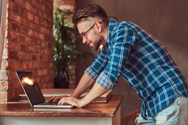 Apuesto estudiante con estilo en una camisa de franela que trabaja en una computadora portátil en una habitación con un interior de loft.