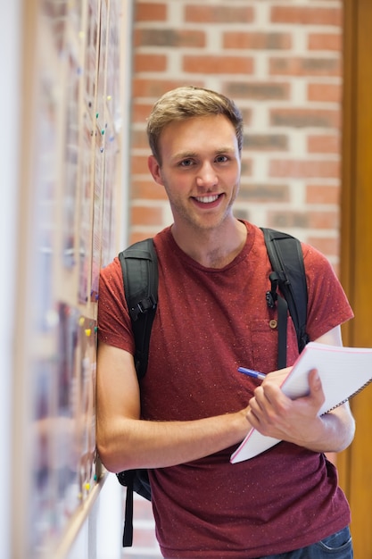 Apuesto estudiante sonriente tomando notas junto al tablón de anuncios