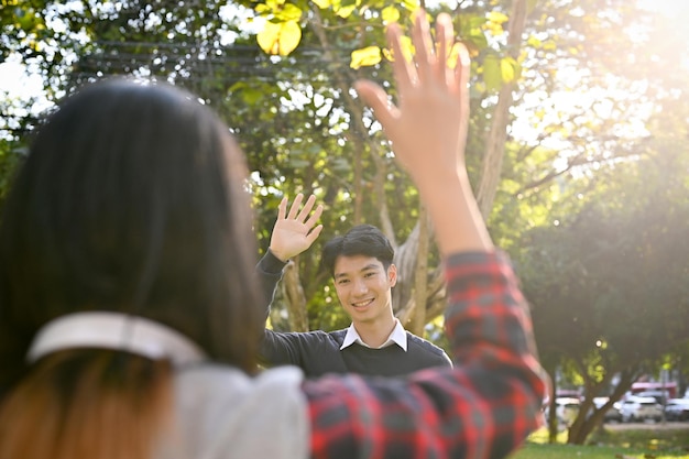 Apuesto estudiante universitario masculino asiático saludando con la mano a su amiga