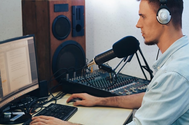 Un apuesto y feliz presentador de radio con auriculares frente a un micrófono y un monitor está grabando un programa de radio en el estudio. Al lado hay una mesa de mezclas y una columna de sonido.