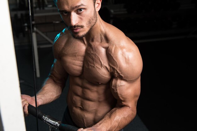 Apuesto fisicoculturista musculoso haciendo ejercicio de peso pesado para bíceps en la máquina con cable en el gimnasio