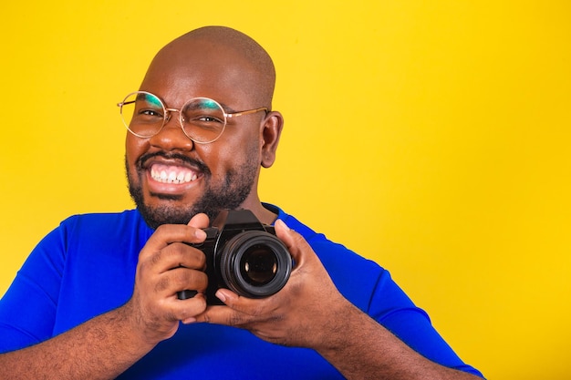 Apuesto hombre afro brasileño con gafas camisa azul sobre fondo amarillo con cámara de fotos fotógrafo sonriente DSLR creativo Foto de primer plano