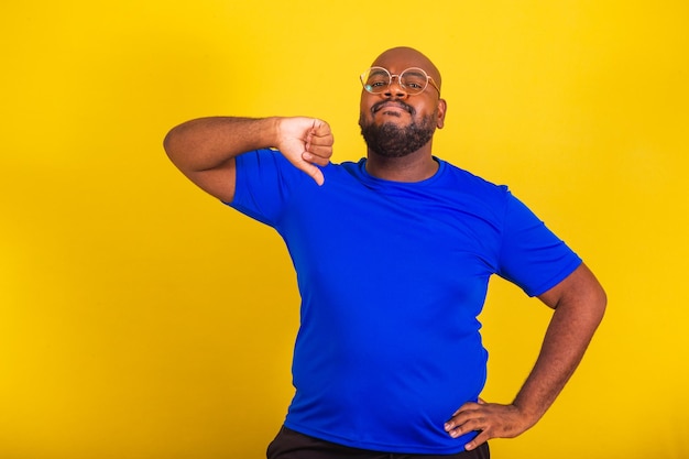 Apuesto hombre afro brasileño con gafas camisa azul sobre fondo amarillo pulgares hacia abajo desaprobando la decepción decepcionado desaprobando negativo