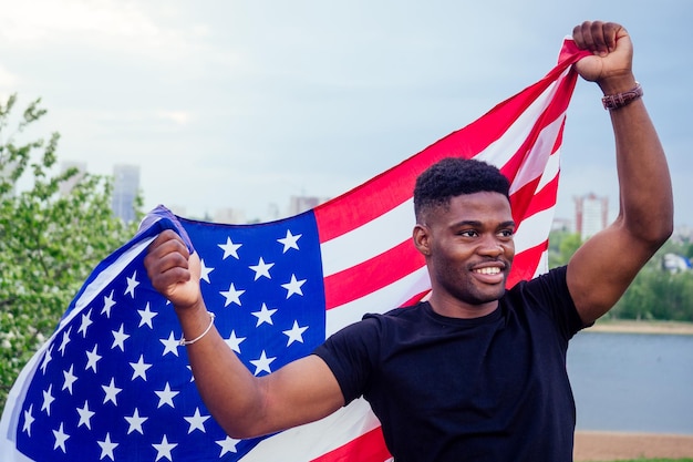 Apuesto hombre afroamericano con la bandera de los Estados Unidos en las manos en el parque de verano cerca de la noche del lago