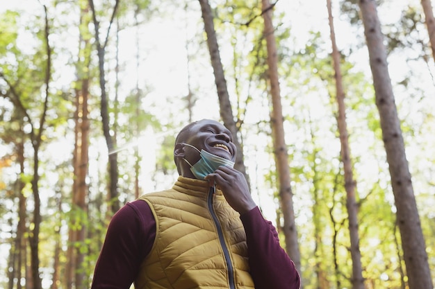 Apuesto hombre afroamericano con ropa informal y máscara médica sonriendo feliz caminando en el parque de la ciudad copia espacio final de la pandemia