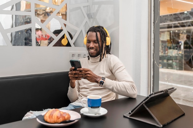 Un apuesto hombre afroamericano sentado en la cafetería usando un teléfono móvil y escuchando música con auriculares amarillos