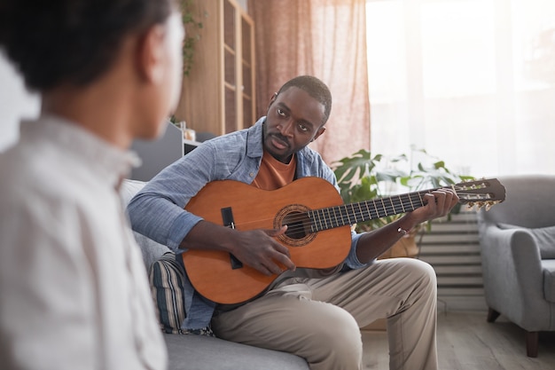 Apuesto hombre afroamericano sentado en el sofá frente a su hijo tocando la guitarra en casa, espacio de copia