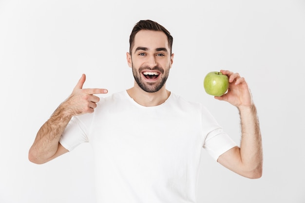 Apuesto hombre alegre vestido con camiseta en blanco que se encuentran aisladas sobre la pared blanca, mostrando manzana verde, apuntando