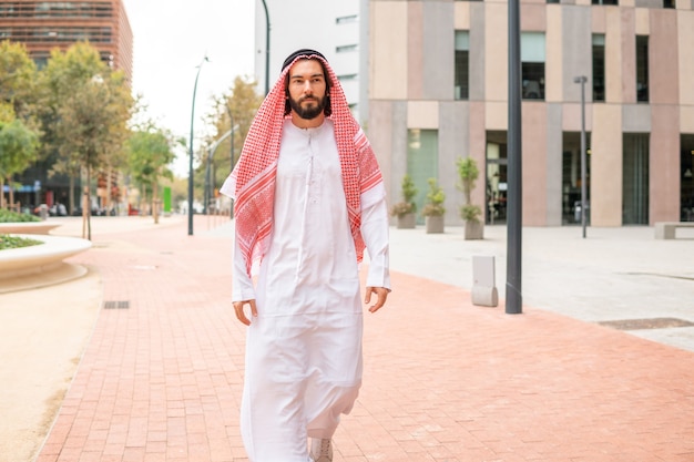 Apuesto hombre árabe exitoso jeque vistiendo ropas tradicionales caminando por las calles de la ciudad en dubai