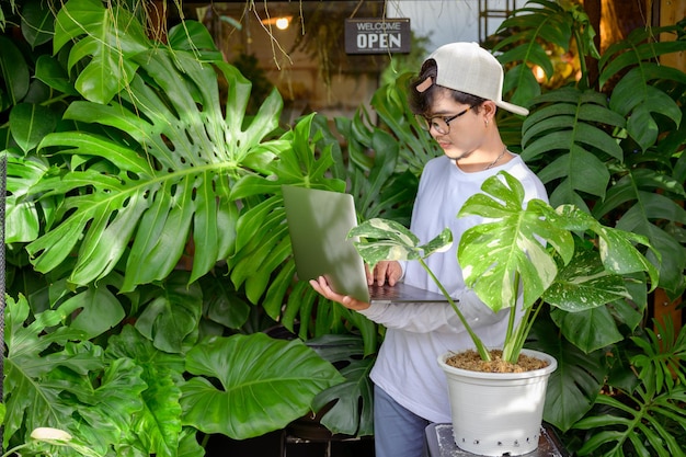 Foto apuesto hombre asiático le encanta plantar árboles en un jardín monstera está usando su computadora portátil para comercializar y vender árboles apuesto hombre asiático está interesado en hacer negocios vendiendo árboles en línea.