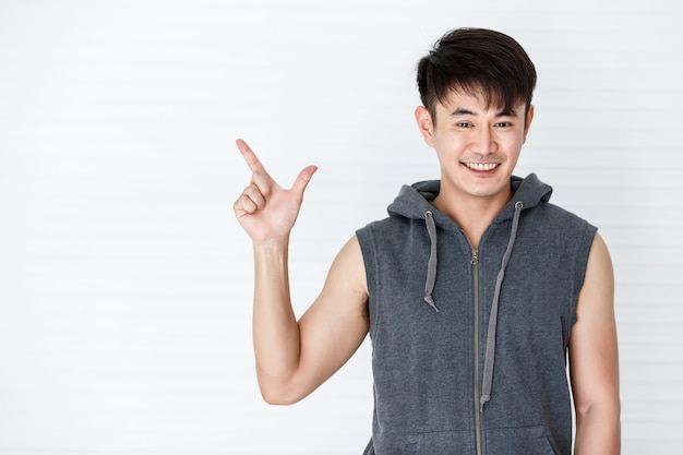 Apuesto hombre asiático en forma sonriente sosteniendo señalar con el dedo para copiar el espacio vistiendo ropa deportiva gris camiseta sin mangas en fondo blanco.