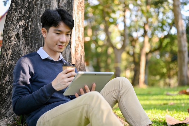 Apuesto hombre asiático usando su tableta portátil mientras se sienta bajo el árbol en el parque del campus