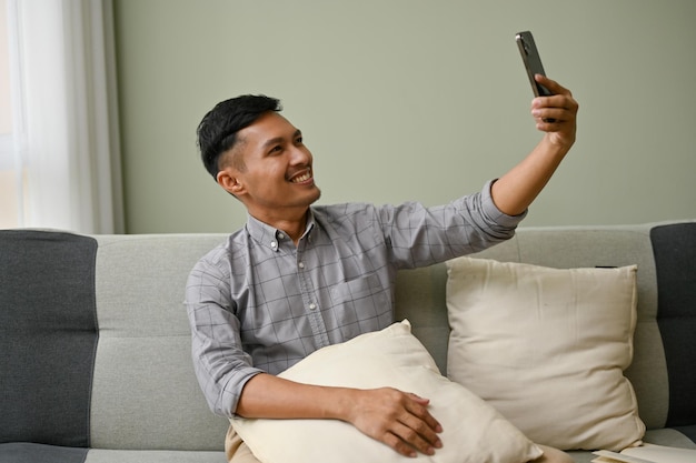 Un apuesto hombre asiático usando su teléfono para tomarse un selfie o un video mientras se sienta en el sofá de la sala de estar