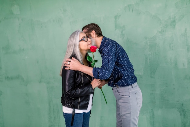 Apuesto hombre barbudo anciano dando hermosa rosa roja fresca a su encantadora esposa sonriente y besando su mejilla, de pie sobre el fondo verde de la pared