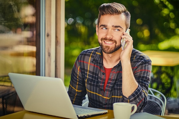 Un apuesto hombre barbudo bebe café mientras usa una laptop en un café.