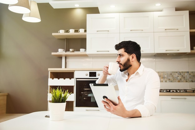 Un apuesto hombre barbudo con el pelo ondulado en la espalda usando una tableta mientras toma un café en una cocina moderna y espaciosa con una elegante camisa blanca